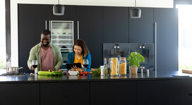 Happy diverse couple cooking, drinking wine and using tablet in modern kitchen, copy space
