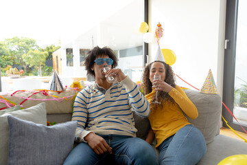 Happy biracial couple having party at home, sitting on couch drinking champagne, copy space