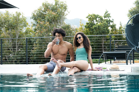 Happy biracial couple relaxing with drinks sitting by pool at holiday home, copy space