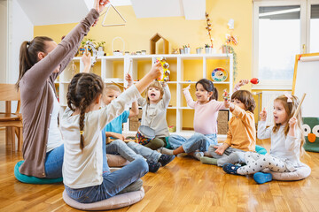 Kindergarten teacher with children sitting on the floor having music class, using various instruments and percussion. Early music education - obrazy, fototapety, plakaty