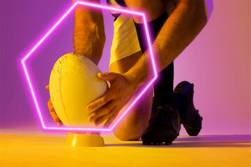 Illuminated hexagon over low section of caucasian male rugby player placing ball on stand - Powered by Adobe