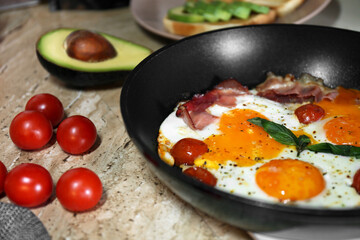 Fried with eggs, bacon and tomatoes in frying pan near ingredients on wooden table, closeup. Tasty breakfast