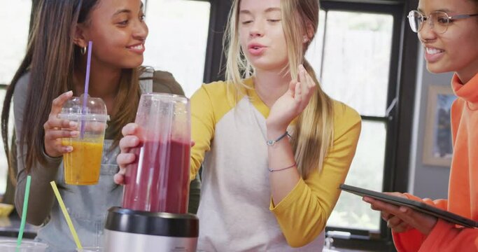 Happy diverse teenager girls friends preparing healthy drink using tablet in kitchen, slow motion