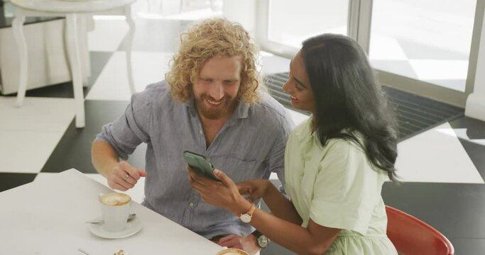Happy diverse couple with coffee using smartphone and talking at a table in cafe