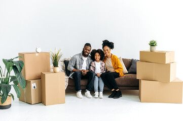 Photo of a happy African American family, joyful dad mom and their preschool daughter, sit on the sofa in the living room between boxes with things, moved to new housing, look at the camera, smile
