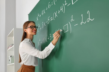 Young math’s teacher writing mathematical equations near chalkboard in classroom