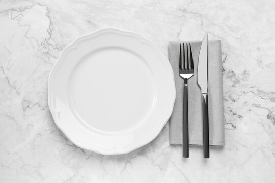 Clean plate and shiny silver cutlery on white marble table, flat lay