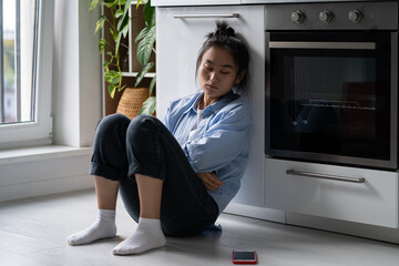 Offended unhappy Asian woman sadly looking at phone sits on floor at kitchen. Sad thoughtful...