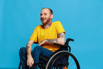 A man in a wheelchair smile looks at the camera in a t-shirt with tattoos on his arms sits on a blue studio background, a full life, a real person