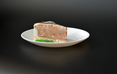 White ceramic plate with a piece of fresh cheesecake decorated with mint leaves on a black background.