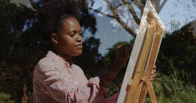 Animation of trees over african american woman painting on easel in garden