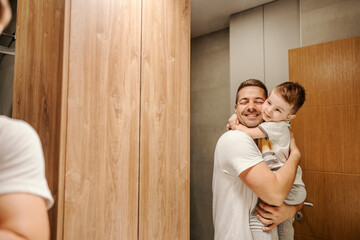 A caring father is holding his son in the arms and hugging him while standing in the bathroom.