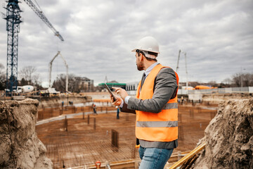 A builder is standing on construction site and checking on works on tablet. He is looking at building foundation.