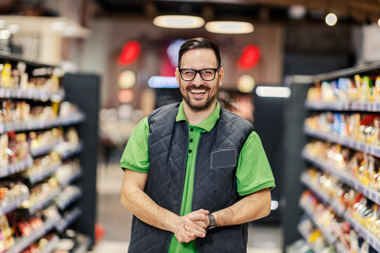 A serviceable salesman is standing at supermarket and smiling at the camera.