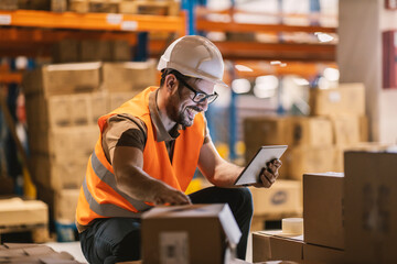 Happy delivery worker is tracking inventory on tablet while crouching in distribution center.