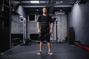 Fototapeta na wymiar A confident male instructor dressed in a special suit for EMS technology stands in the gym with a dark gray atmosphere. Revolution in training, body rehabilitation, fitness workers and staff