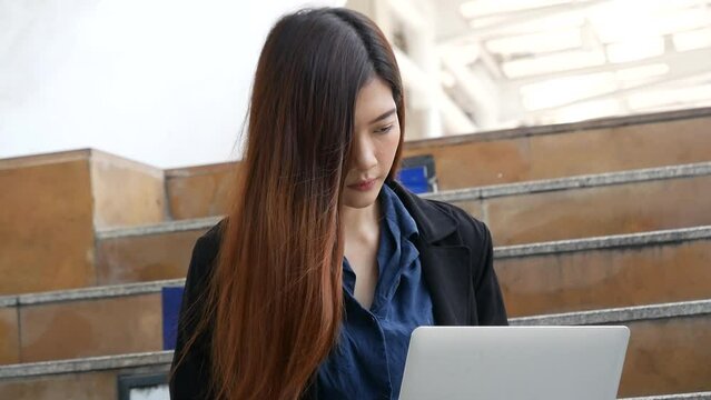 Businesswoman using laptop typing keyboard drinking coffee cup with internet technology lifestyle. Asian woman use cellphone sit modern city street outdoor. Smart woman use laptop work from anywhere