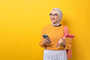 Happy young Asian student girl with backpack holding notebook and mobile phone, looking away at copy space isolated over yellow background