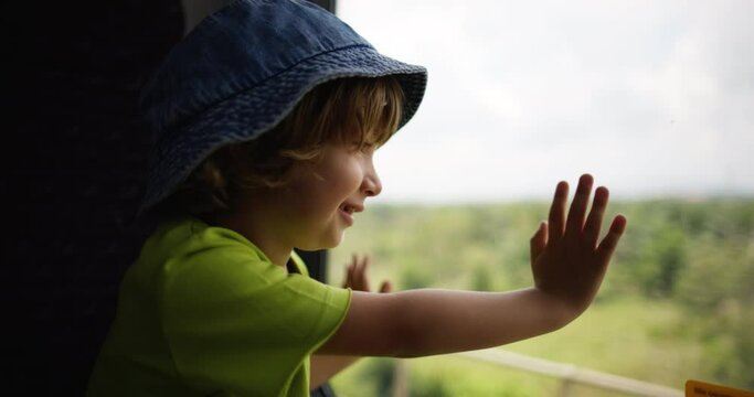 A joyful and happy boy in panama hat leaned his palms on glass of the train and looks out the window, following the changing views. The child boy smiles and looks enthusiastically out the train window