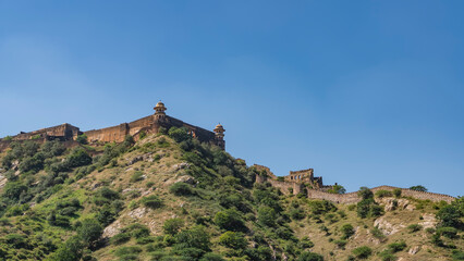Fototapeta na wymiar An ancient defensive fortress wall runs along the ridge of the mountain. Watchtowers with domes are visible against the blue sky. Green vegetation on the slopes. India. Amber Fort. Jaipur