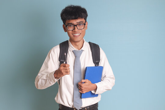 Indonesian senior high school student wearing white shirt uniform with gray tie holding some books, smiling and looking at camera. Isolated image on blue background