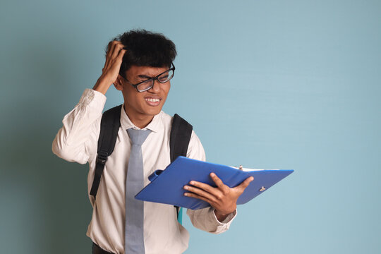 Indonesian senior high school student wearing white shirt uniform with gray tie writing on note book using pen and thinking about an idea. Isolated image on blue background