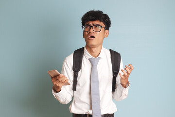 Indonesian senior high school student wearing white shirt uniform with gray tie feeling disappointed making angry hand gesture while holding mobile phone. Isolated image on blue background