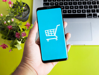 hand holding smart phone with shopping cart logo on screen along side with flower and laptop on yellow background