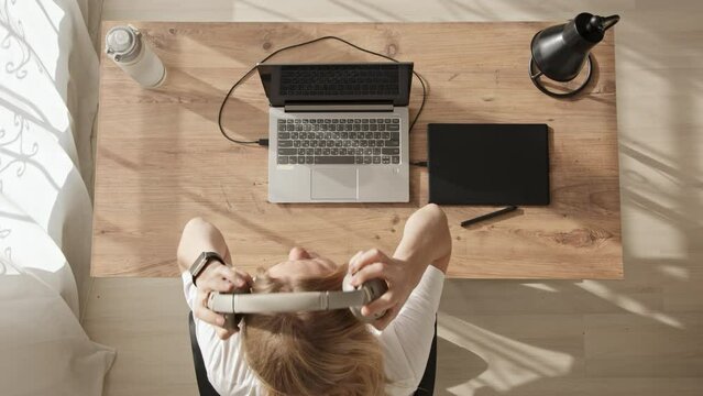 Top down view of girl at desktop with laptop and graphics tablet. Young blonde girl draws sketches or illustrations on black graphics tablet at home. Freelance work during day in bright room.