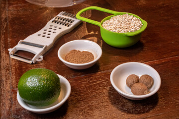 ingredientes da receita separados em tigelas com limão, noz-moscada, canela em pó, uma panela verde com aveia em flocos e um ralador sobre uma mesa rústica de madeira.