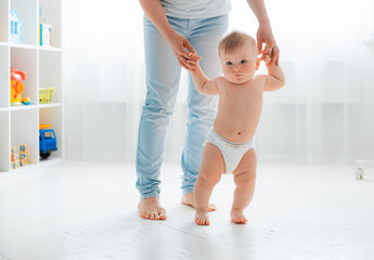 Cute little baby in a diaper learning to walk in a white room , mom holding his hands