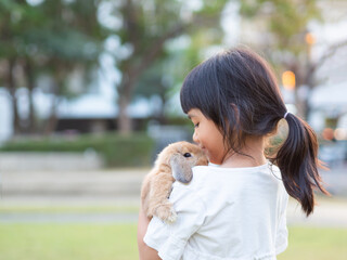 Kind little cute girl 7 years old holding a baby holland lop rabbit on shoulder at the garden. Child with a gentle heart, loves animals.