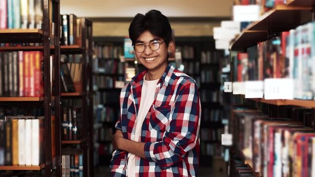 Asian man in glasses and a plaid shirt stands in library smiles looking at camera. An Oriental exchange student man stands between the shelves of books the archive and looks at camera smiling