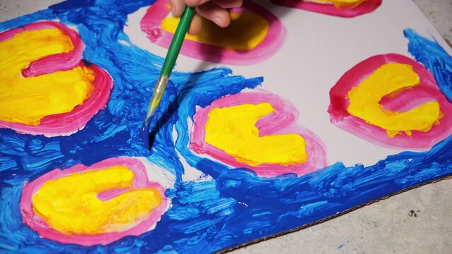 Child is painting colorful hearts by brush and blue, yellow, red colors gouache paint on cardboard . Boy is drawing the Mothers day card or fathers day. Creative Artistic hobby. Love dreams. Top view.