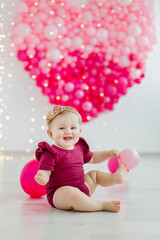 A happy smiling adorable baby girl in a burgundy red romper is sitting on the floor with balloons in St. valentine's day decorations. Big heart made of pink balloons in the background. 