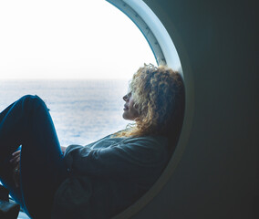 Serene woman sitting inside a porthole in the boat cruise ferry transport sea looking the ocean water and enjoying the trip alone. Passenger people dreaming destination. Relax and journey lifestyle