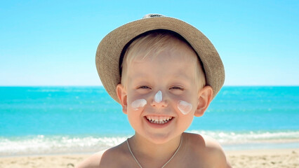 Positive emotional happy kid, smiling and laughing in a trendy hat, sunscreen on his face against the backdrop of sea waves. Rest, swim on the paradise ocean beach. Vacation and holidays with family
