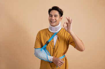 Handsome young asian man with broken arm in soft splint suffering a sore arm showing ok sign isolated on beige background, accident insurance concept. - 566919546
