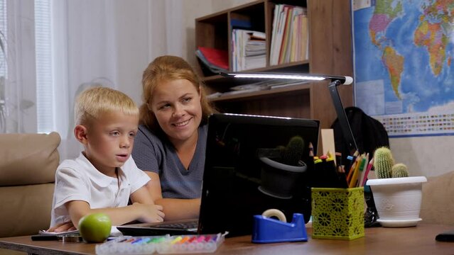 A smiling adult mother and her young son are holding a video conference with a teacher during distance learning, they are sitting at a desk at home against the background of bookshelves.