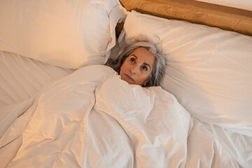 An old gray-haired woman lying on a pillow in bed woke up at night looking at the ceiling and tries...