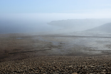 Amazing lonely lake shore in early morning with soft haze above smooth tranquil water with pebble coast and cape in fog. Liveless and unearthly landscape.