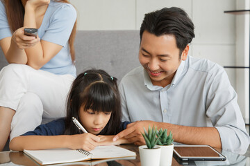 Obraz na płótnie Canvas Children education and home school concept : Young asian father and mother see little daughters' study. Excited smiling small child girl enjoying learning and writing with pleasant dad and mom at home