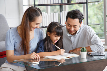 Children education and home school concept : Young asian father and mother see little daughters' study. Excited smiling small child girl enjoying learning and writing with pleasant dad and mom at home