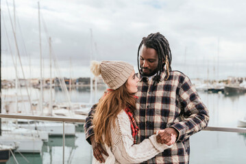 Fototapeta premium a multiethnic couple looking at each other holding hands, the harbor in the background