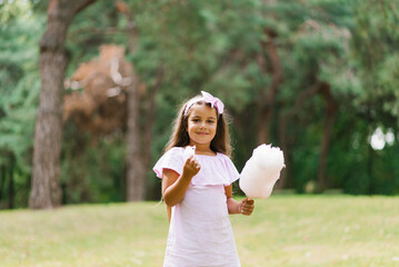 Adorable and pretty girl eating white sweet cotton candy. Happy child, kid eating sweets with emotions in the park at summer