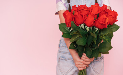 Little girl in denim overalls holding bouquet of roses behind her back against pink background. mother's day