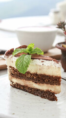 Delicious Tiramisu Cake is a typical Italian cheese cake topped with cocoa powder and mint leaves as decoration served on a white plate with a fork.