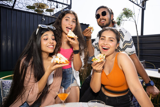 Group of cheerful indian friends having fun eating pizza talking and laughing together while drinking cocktails at restaurant cafe. Summer season, food and drinks. People enjoy vacation and party