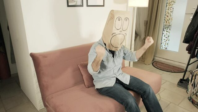 A man, wearing a funny breadbag face on the head, watching the tv on the sofa at home, sharing happiness with someone else in the room by rasing his hands.
