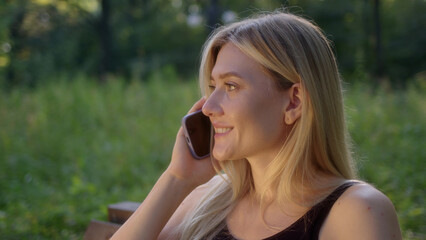 Attractive young woman talking on phone in park. blonde in black sports top sits on bench and chats on smartphone. girl is glad to hear answer and smile.